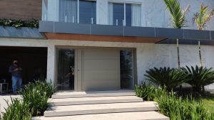 Porta pivotante em ACM Silver Gold com frisos horizontais, puxador cava, portal duplo lateral com vidro 10 mm incolor e dupla vedação