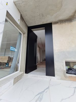 Porta pivotante em ACM preto carbono, painel lateral fixo no mesmo alinhamento da porta, puxador exposto de inox com 2 metros cromado e dupla vedação - Imagem 6