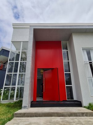 Porta pivotante em ACM com duas cores. Lado externo vermelho fosco e lado interno cinza. Painéis e bandeira fixos seguindo mesmo alinhamento da porta e dupla vedação. - Imagem 2
