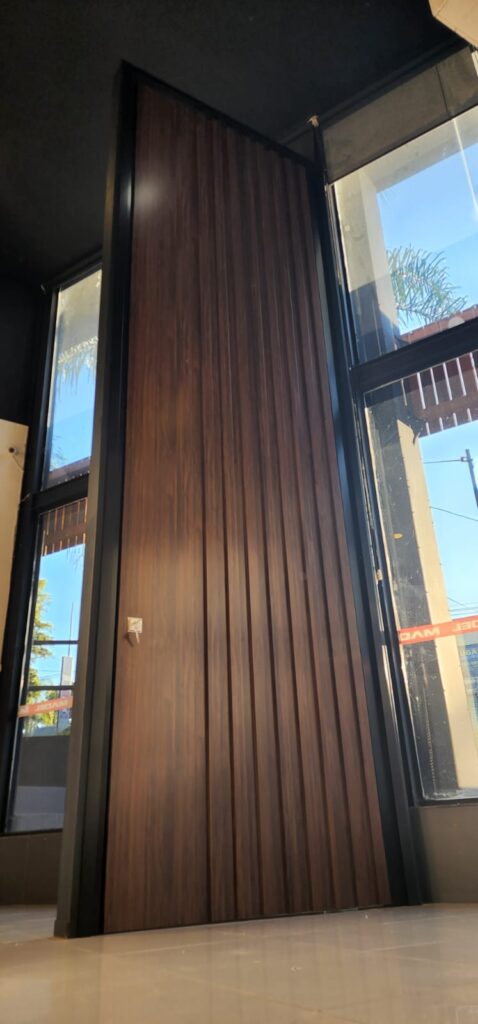 Porta pivotante em ACM ripado na vertical cor Sucupira e dupla vedação ...
